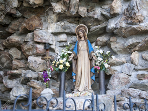 A statue of the Virgin Mary stands adorned with flowers within a natural rock alcove. She is clothed in a blue robe over a beige dress, hands clasped in prayer. A small cross is to her left. The setting appears serene and rustic with a clear focus on the spiritual symbol.