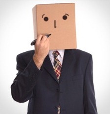 A person wearing a suit with a cardboard box on their head, featuring drawn-on eyes and mouth, holds a marker to the box as if drawing the mouth.