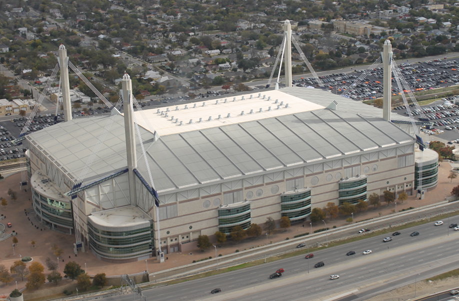 Aerial view of a large stadium with a white retractable roof supported by long white pillars, surrounded by parking lots and located next to a busy highway.