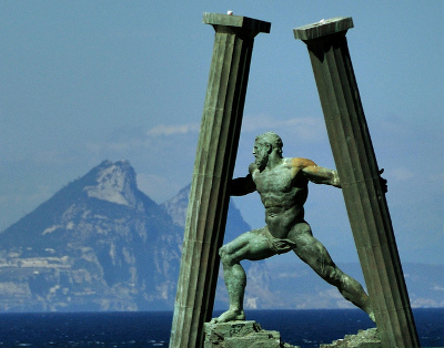 A bronze statue of a muscular man posed dynamically, positioned between two tall, ancient stone columns. In the background, a prominent mountain looms over a calm sea under a clear blue sky.