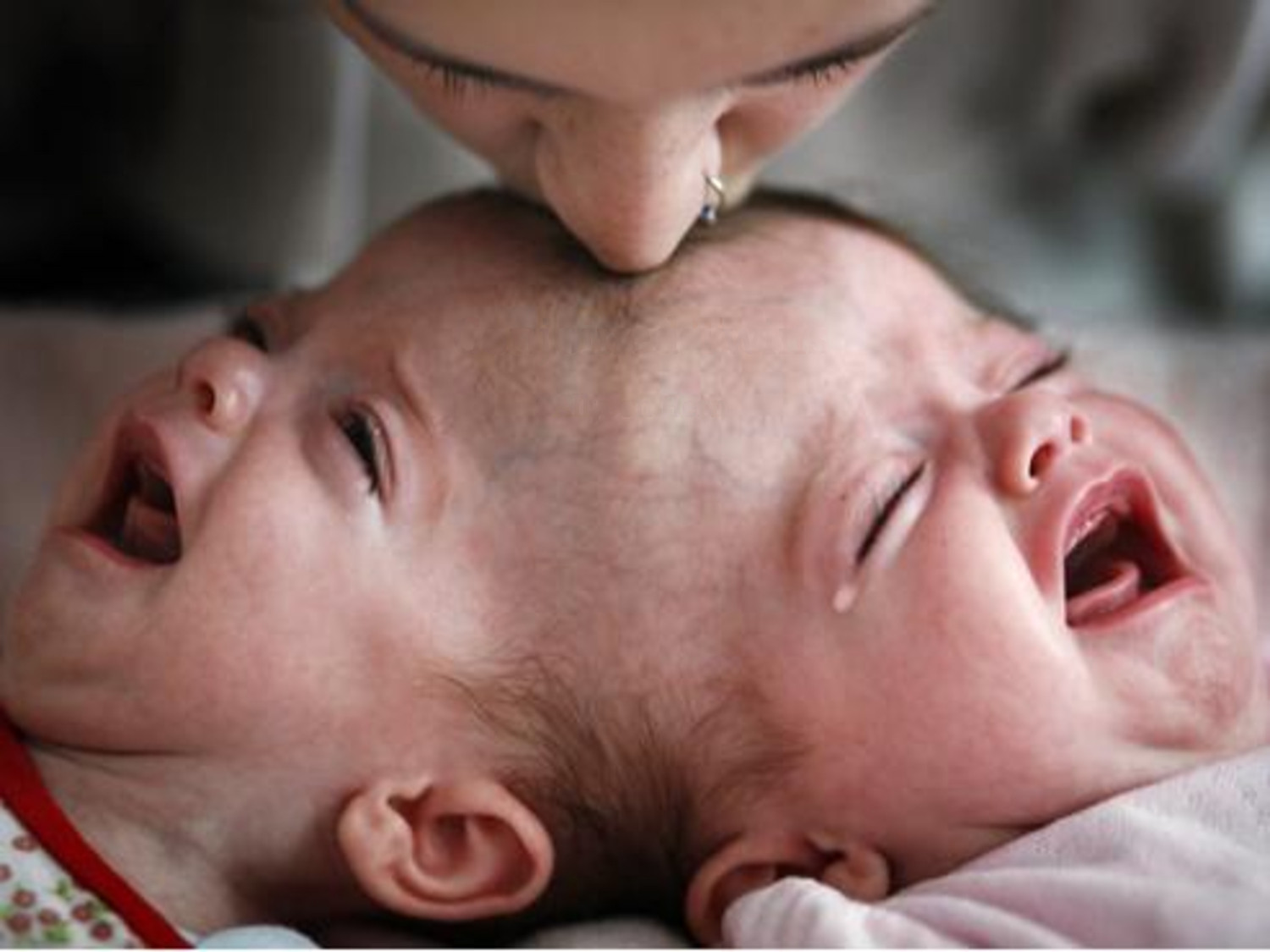 Image showing a tender moment between an adult and two crying infants. The adult, whose forehead is visible, gently kisses one of the babies on the head, who appears distressed.