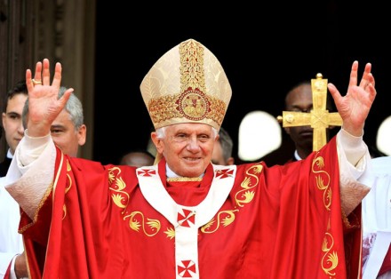 A senior religious leader in red and gold vestments, with a mitre, raises his arms in a greeting or blessing gesture, surrounded by younger aides in white robes.