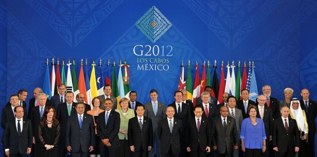 A group of global leaders from various countries standing in two rows in a formal setting at the G2012 summit in Los Cabos, Mexico, with a backdrop displaying the event's logo and multiple national flags.