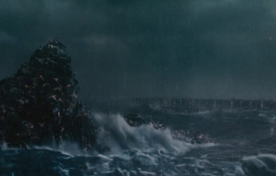 A tumultuous sea under dark, stormy skies crashes against a rocky outcrop, with a faint silhouette of a distant bridge in the background.