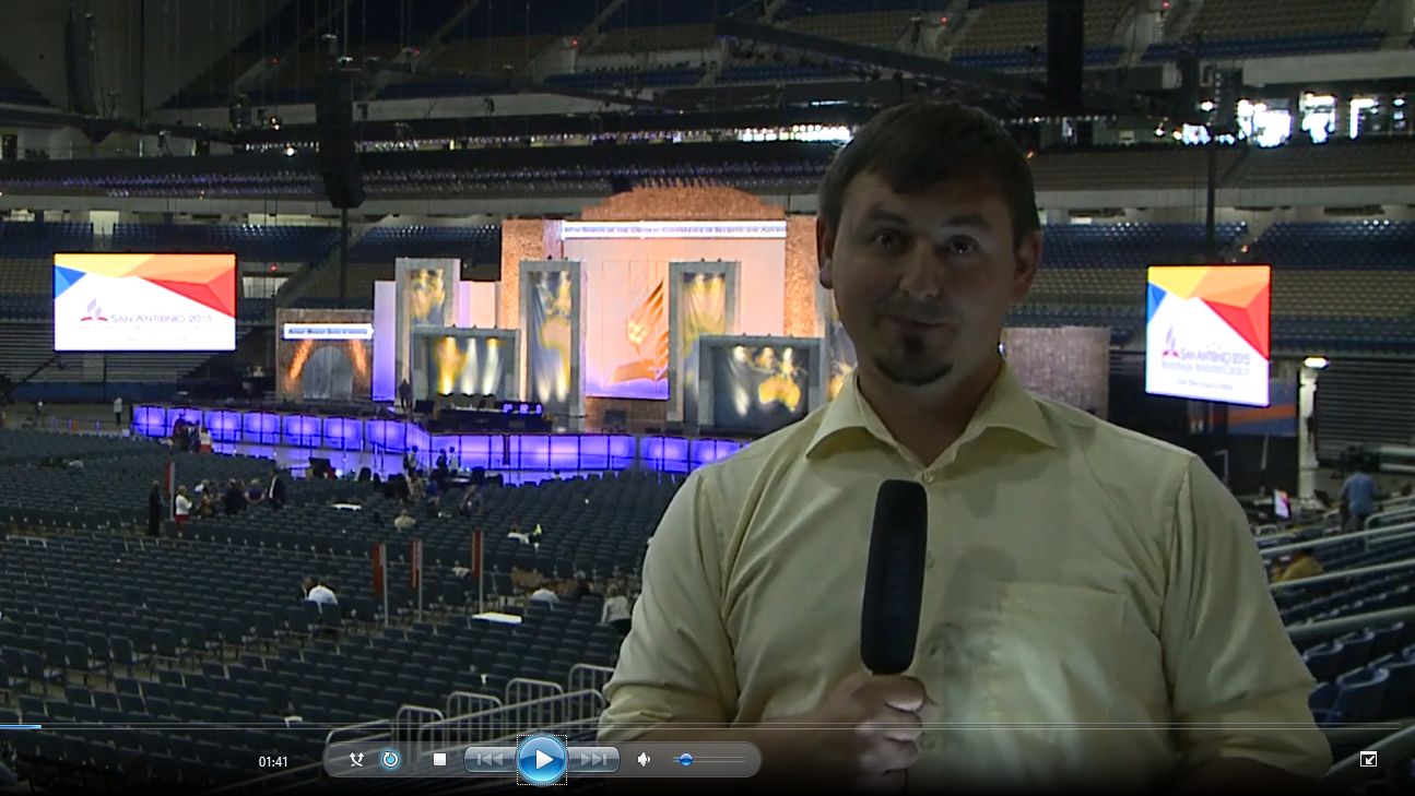 A man in a light yellow shirt holding a microphone stands in the foreground, with a large stadium event setup in the background featuring screens displaying images and logos related to a 2015 event in San Antonio.