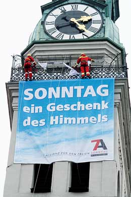 A tall clock tower shows Roman numerals and hands pointing to three o'clock. Two figures dressed as Santa Claus decorate the top, spreading a banner below the clock that reads "SONNTAG ein Geschenk des Himmels," translating as "Sunday, a gift from the heavens." The scene underlines celestial celebration themes rooted in traditional belief systems.