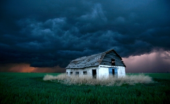 An abandoned white house with a deteriorating roof sits isolated in a vast field of green grass under a dramatic stormy sky showing dark clouds and subtle hints of lightning in the distance.