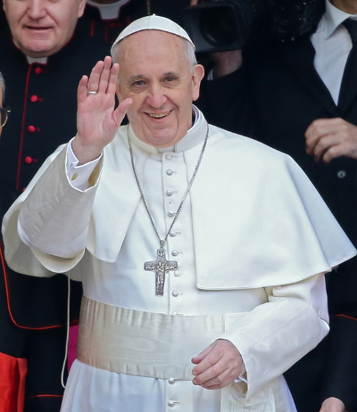 A man in elaborate white religious attire, sporting a prominent silver cross, waves cheerfully amidst a blurred background of spectators.