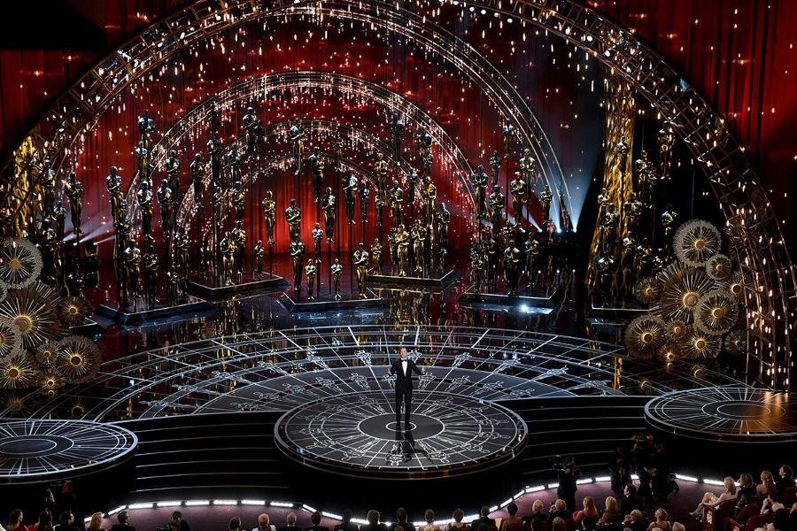 An opulent event stage adorned with celestial-inspired motifs including arcs and circles, resembling an observatory under a universe-themed dome. Multitudes of golden statues line the stage's perimeter, creating a dramatic and elegant atmosphere. A lone figure stands at the center on a podium, speaking to an attentive audience seated in a darkened area.