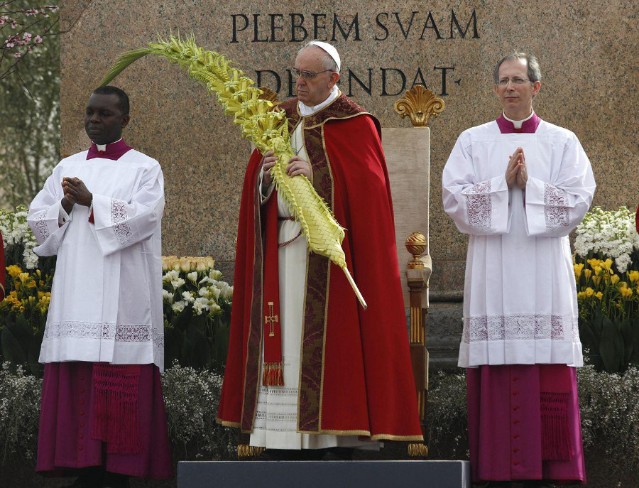 Three religious figures in ceremonial attire stand before a floral backdrop. On the left, a man in a purple cassock and white surplice clasps his hands in prayer. In the center, a man in a red chasuble holds a large palm branch, symbolizing peace and victory. On the right, another man in a white robe and purple stole stands with hands joined in prayer. Behind them, a stone monument adorned with the phrase "PLEBEM SUAM DILEXIT" is visible.