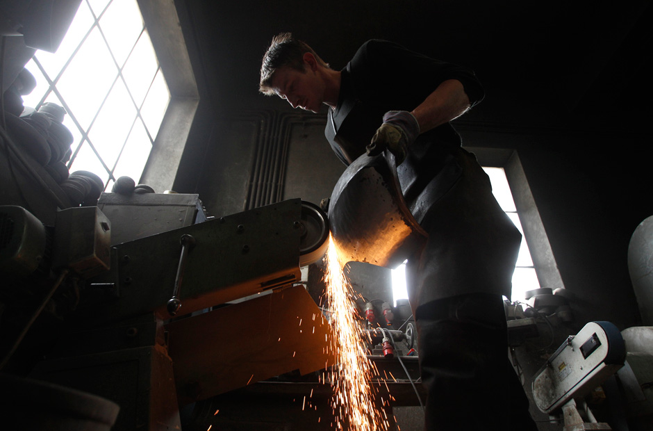 A man in a dark workshop pours molten metal from a crucible, causing sparks to fly. The silhouette of his face and upper body is visible against the backlight from a window.