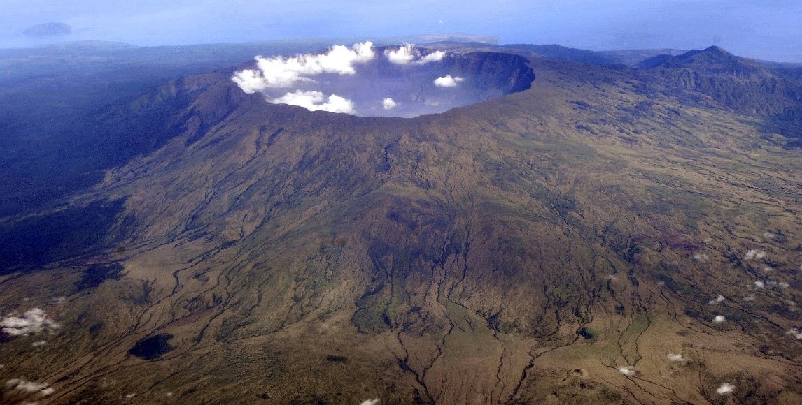 Aerial view of a large volcanic crater surrounded by expansive, rugged terrain with distinctive erosional gullies, under a clear sky with a few scattered clouds.