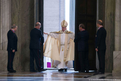 A religious leader dressed in ceremonial robes is surrounded by four men in formal attire inside a large, marbled hall.