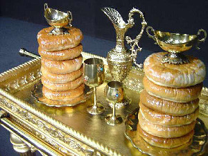 A traditional display features stacks of ring-shaped breads alongside ornate golden cups and a pitcher, arranged on a reflective gold tray against a dark background.