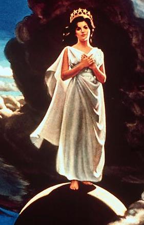 A woman stands on a crescent moon set against a cloudy sky, dressed in a flowing silver robe and wearing a crown. She holds one hand to her chest and looks contemplative.