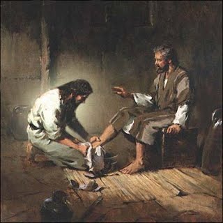 An oil painting depicting two men in a rustic, dimly lit room. One man, with a gentle expression, washes the bare feet of another older man who is seated and gesturing. Both are dressed in traditional Biblical-era garments.