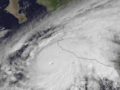 Satellite image showing a powerful hurricane with a distinct eye, approaching a coastline. The swirling cloud formations suggest its immense scale and intensity as it moves over the ocean.