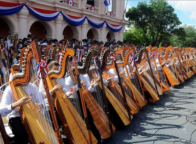 A large group of musicians dressed in white shirts and dark pants, playing harps in front of a festively decorated building with red, white, and blue banners.