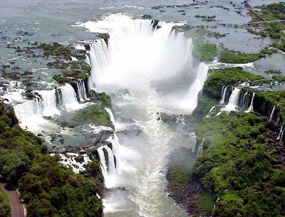 Aerial view of a large, powerful waterfall surrounded by lush greenery, with water cascading forcefully into a river below.