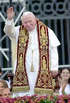 A religious figure dressed in ornate vestments raises his hand in greeting, standing before a blurred backdrop of onlookers.