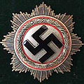 Decorative disk featuring a central black swastika enclosed within a white circle, bordered by a red ring, and surrounded by a radiant silver and gold sunburst pattern on a dark background.